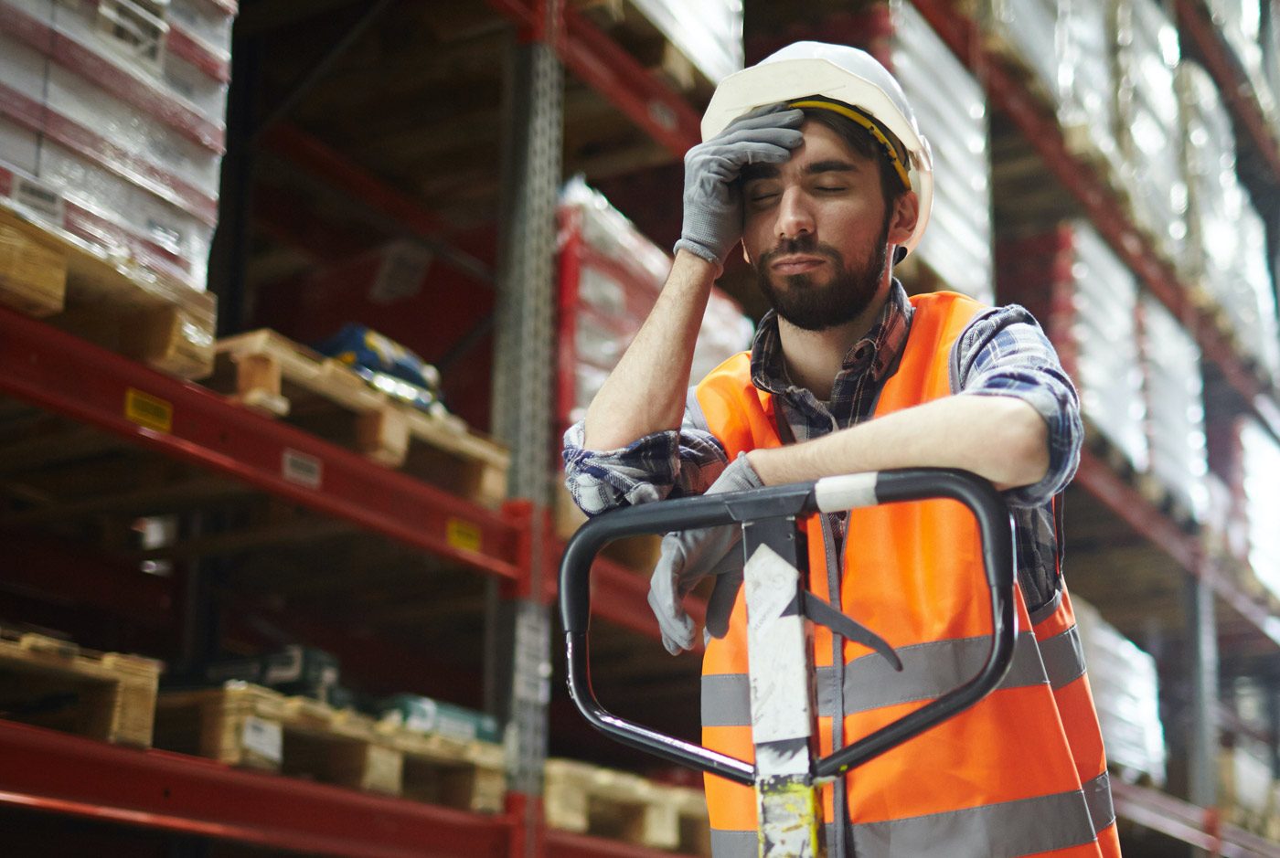 Man wearing a hard hat, safety vest and gloves while falling asleep in a warehouse
