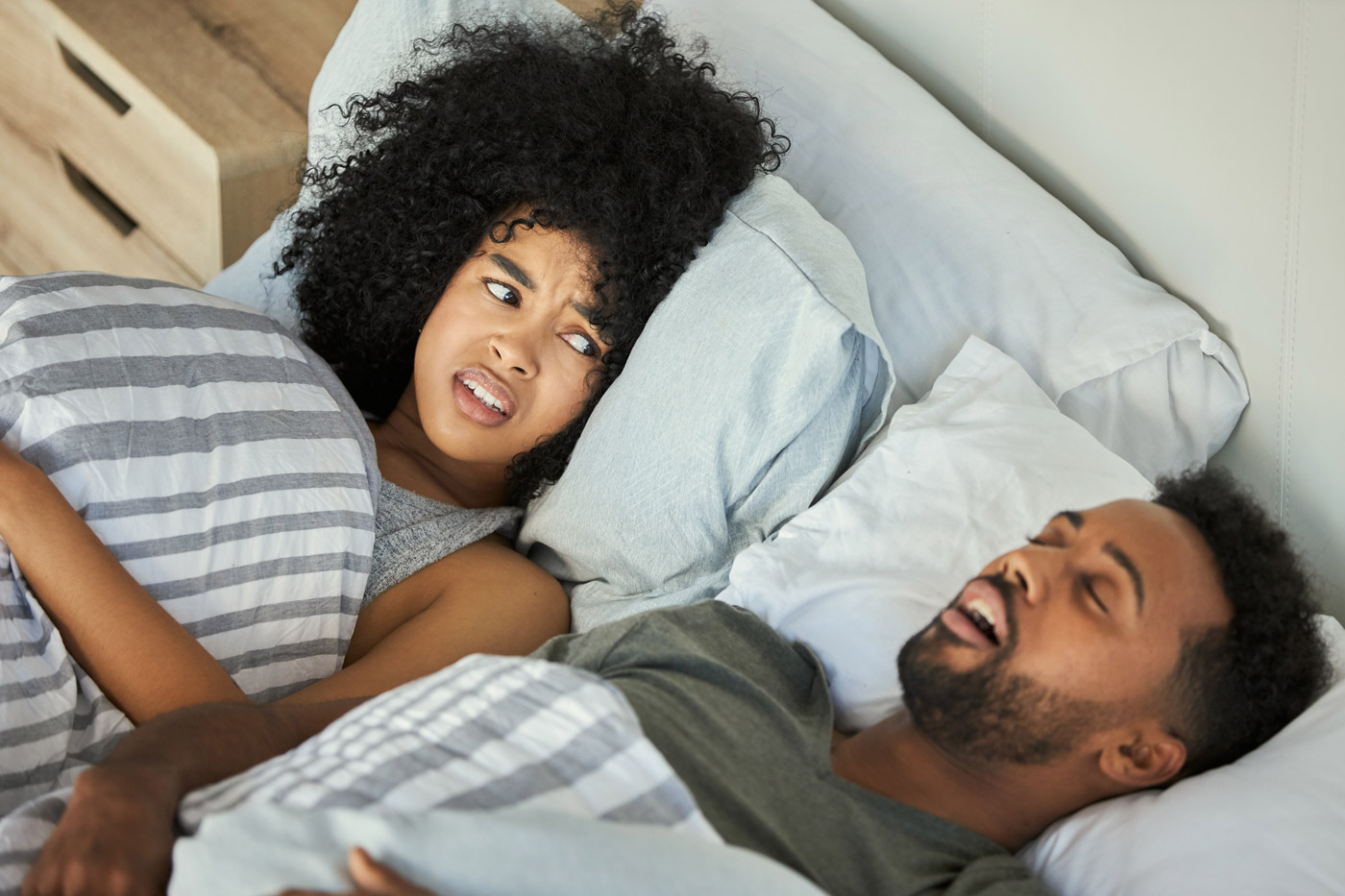 A woman lying in bed looks annoyed at her sleeping partner