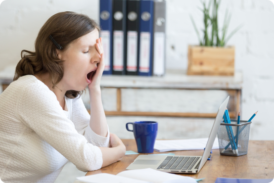 Woman yawning and sitting at her desk in the office with a laptop and coffee