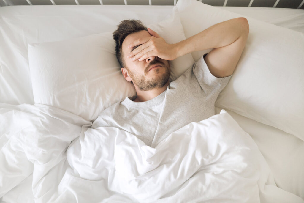 Man laying in white bed sheets with hand over his face - Restless legs syndrome