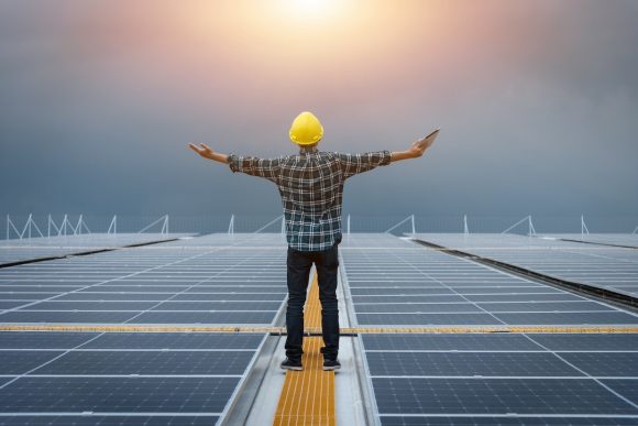 Technician inspecting the installation of solar panels standing with arms outstretched on solar panels against sun sky background, solar power clean energy, concept great power